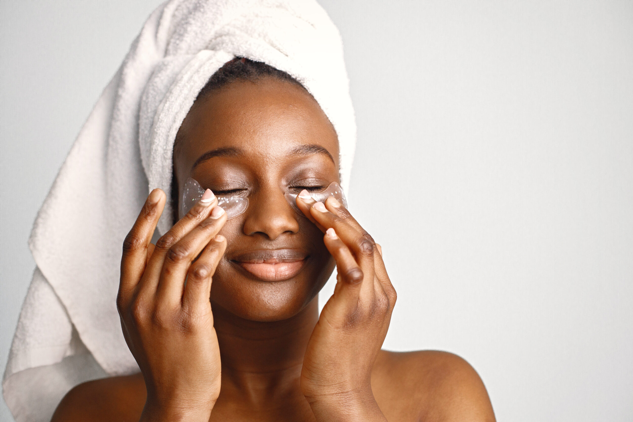 Black young woman standing isolated on white background with patches on her face. Girl has her hair wrapped in a towel. Black  woman enjoy after body treatment.